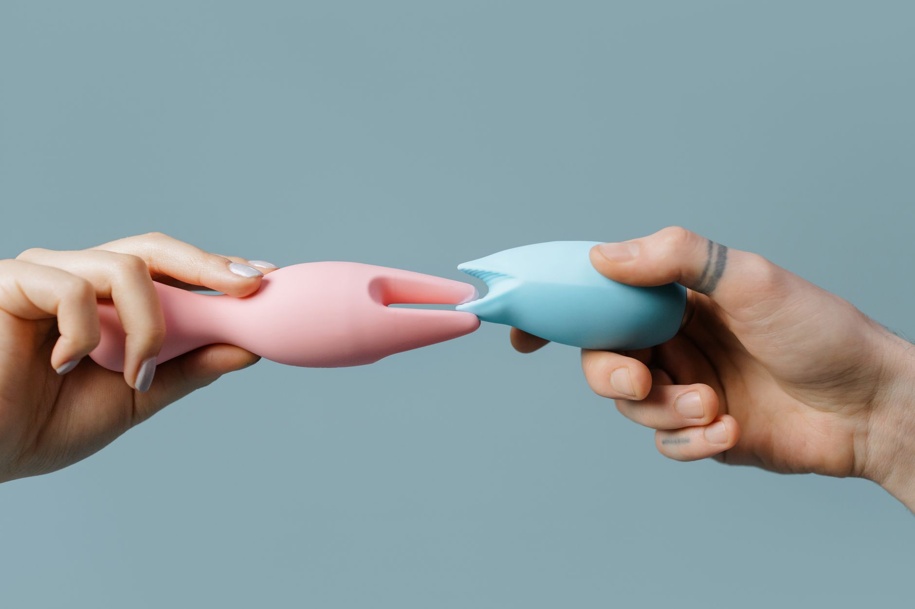 Two hands holding pink and blue silicone vibrators against a pastel background